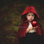 A young girl wearing a red cape offering a juicy red apple