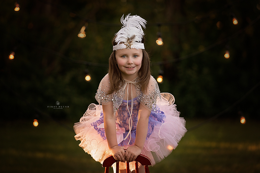 Little girl dressed in a tutu and feather headband stands on a red chair with smiles thanks to Make them smile philosophy.